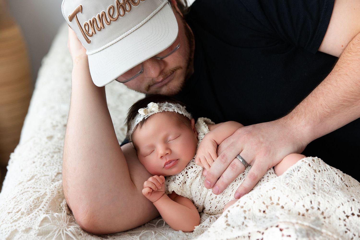 A newborn baby sleeps peacefully on white lace fabric while being gently held by adult hands.