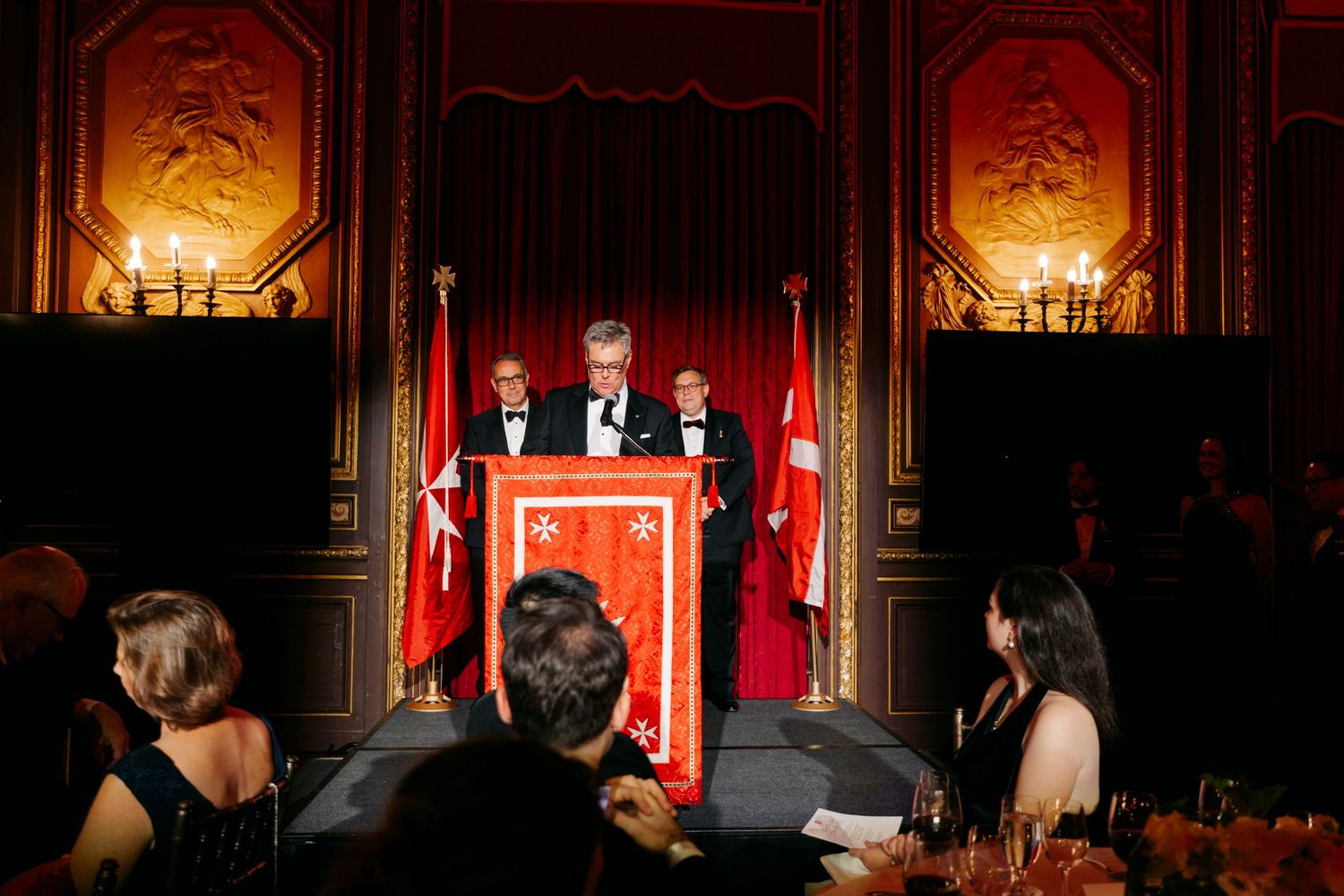 Performers on stage during a theatrical production with ornate red and gold decor in the background.