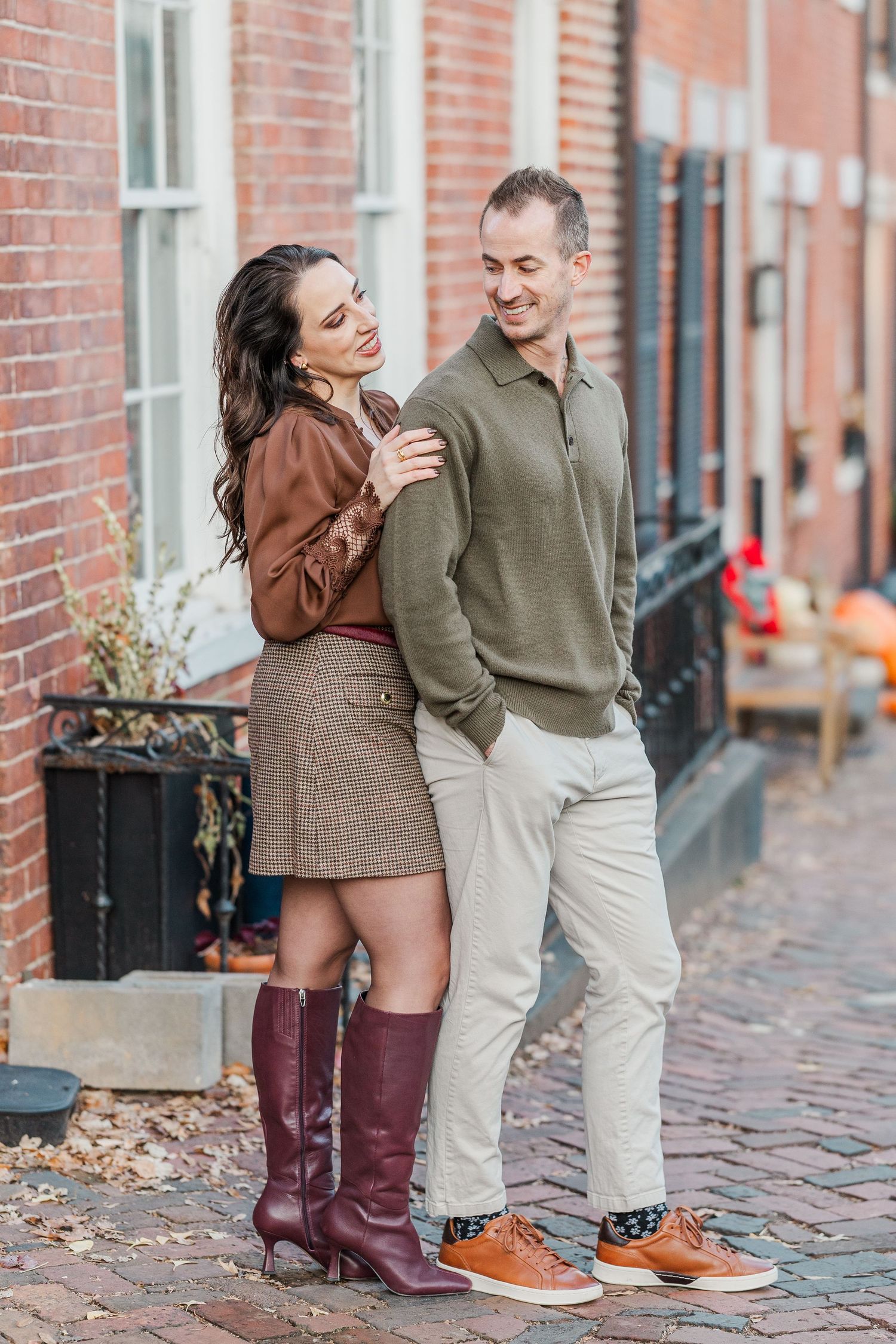 Couple in casual fall outfits sharing a playful moment on a brick sidewalk, showing burgundy boots and orange shoes.