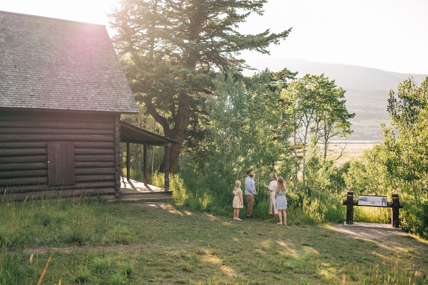 A rustic log cabin sits nestled among green trees and rolling hills during golden hour.