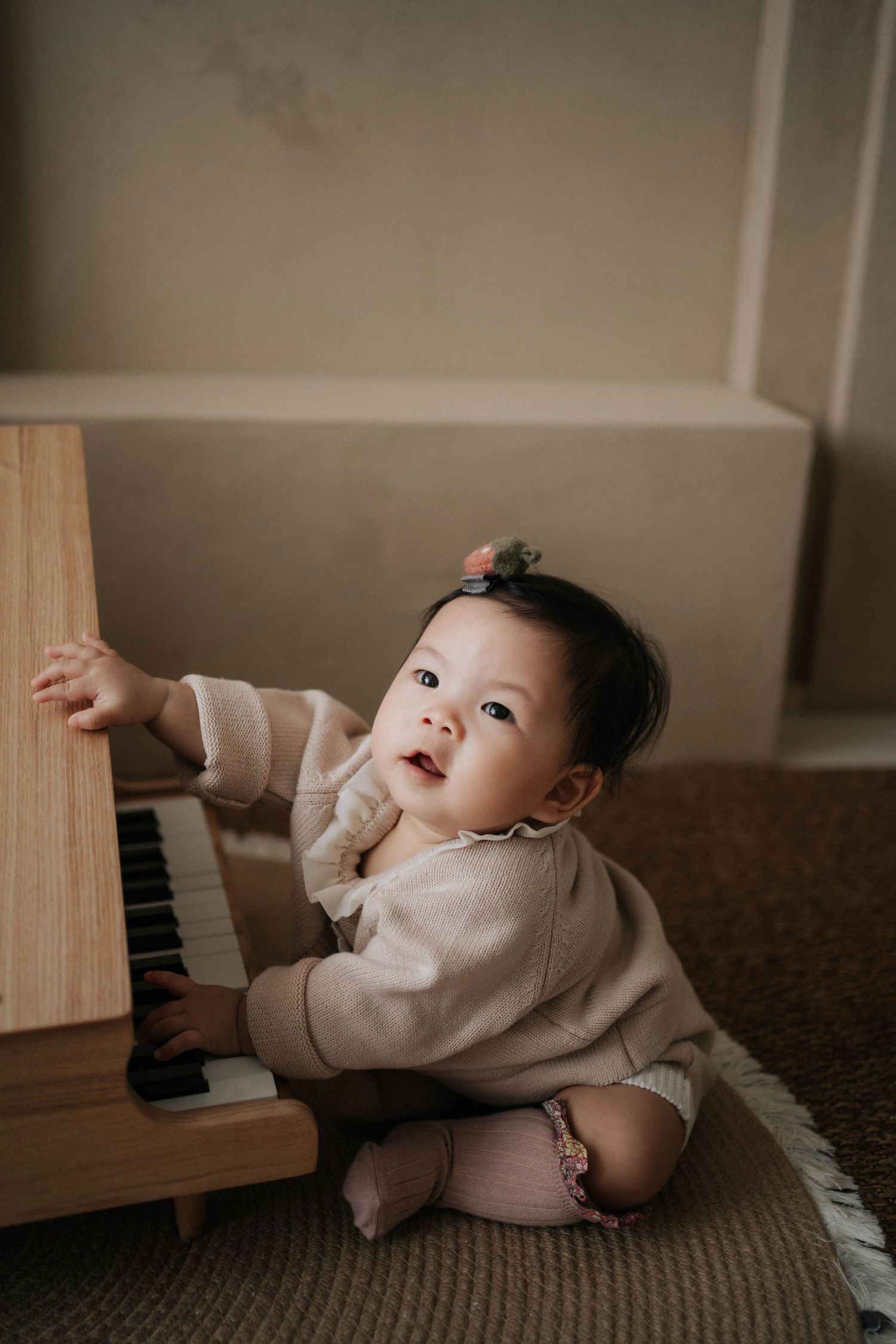 Baby girl in beige outfit sits on floor playing with a small wooden toy piano, looking up curiously.穿著米色衣服的女嬰坐在地板上玩著小型木製玩具鋼琴，好奇地抬頭看著地面。