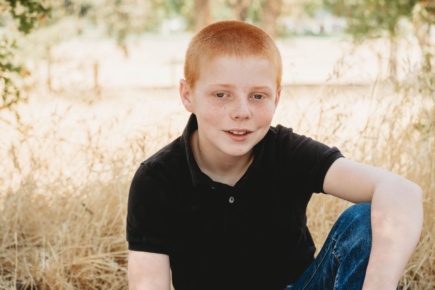 A child in a black shirt sits outdoors among tall golden grass during a sunny autumn photoshoot.