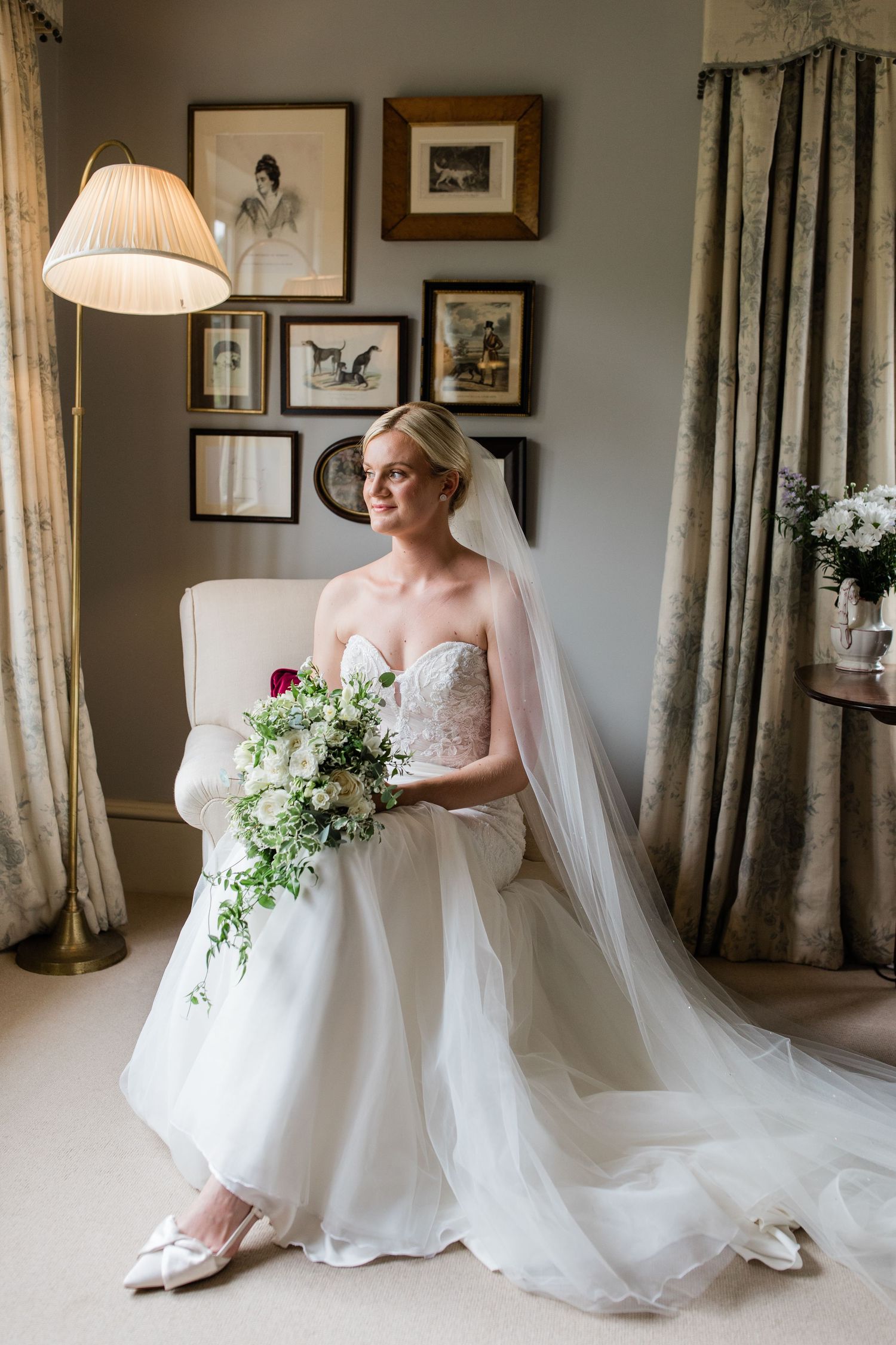A bride in a flowing white wedding gown and veil poses gracefully in an elegantly decorated room with framed artwork.