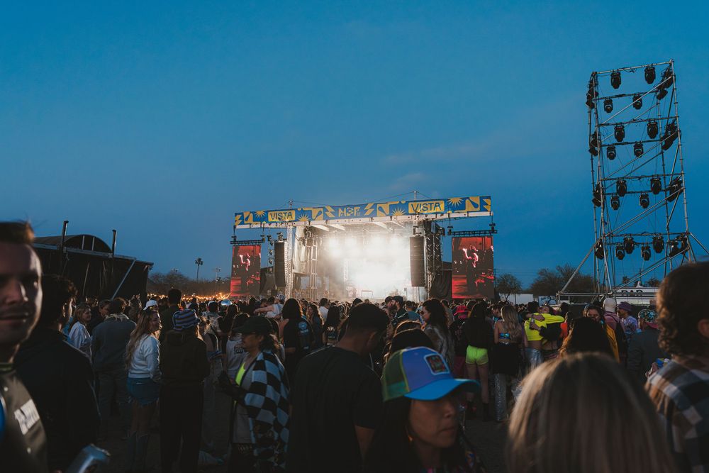 Large crowd of concert attendees watching an outdoor music performance with bright stage lights at dusk.