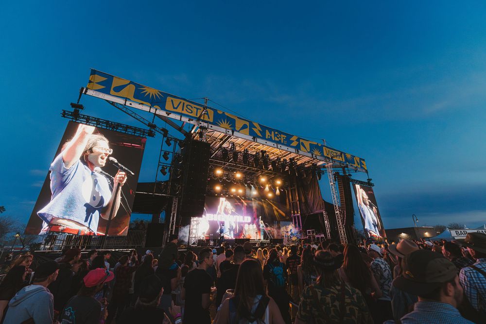 Large outdoor music festival stage illuminated at dusk with crowd silhouettes and vibrant blue sky backdrop.