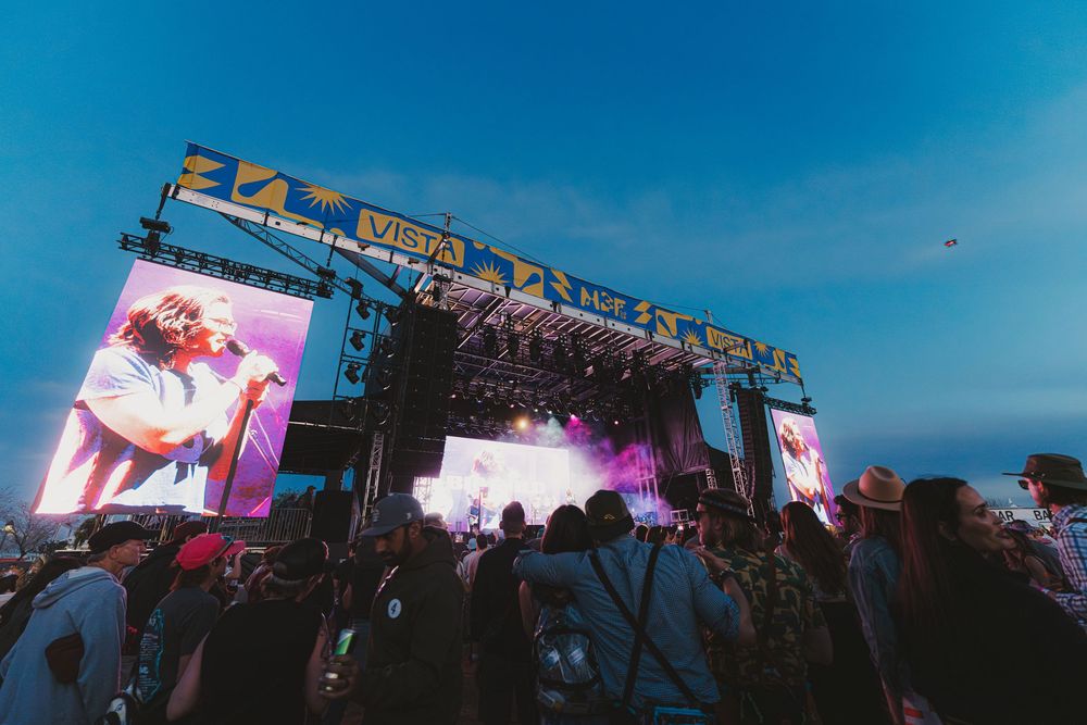 Concert stage with large video screens and lighting displays glows against a vibrant blue sky at an outdoor music festival.