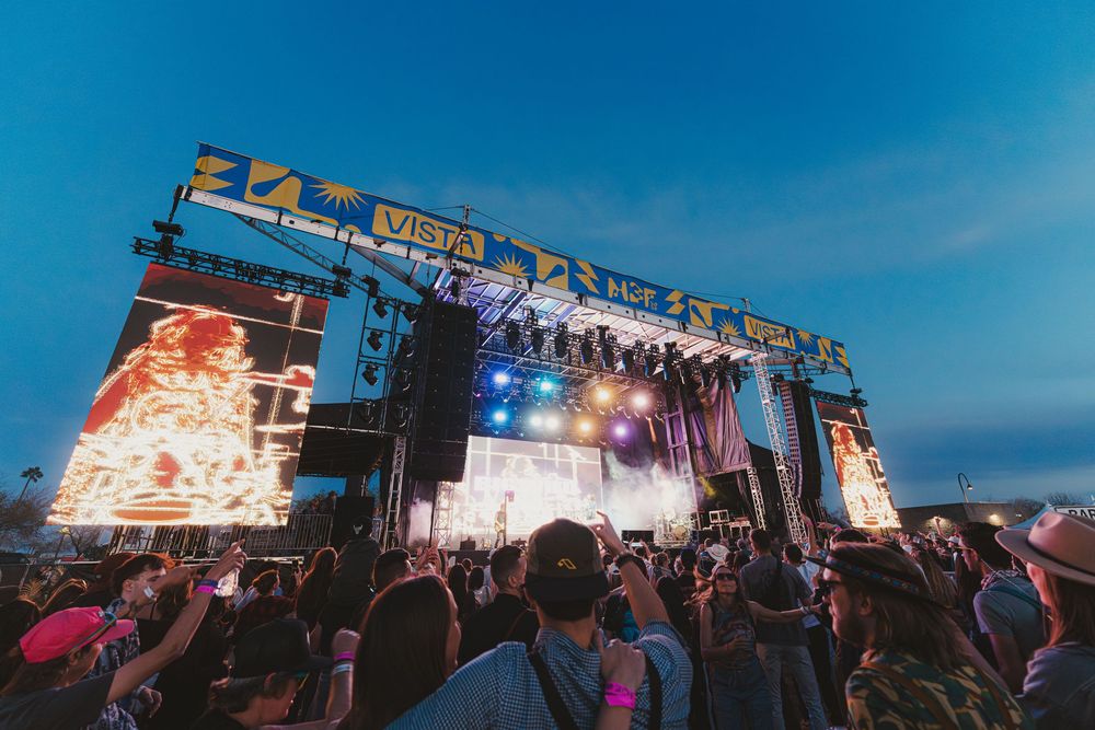 Outdoor music festival crowd enjoying a concert under blue sky with stage lights and equipment visible.