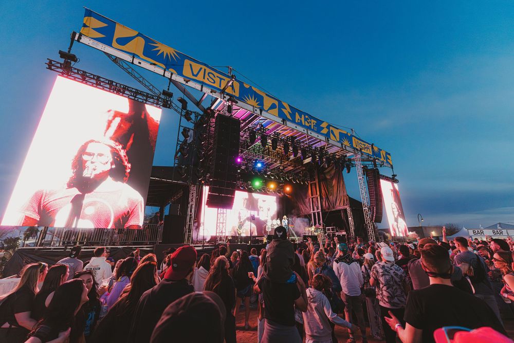 Large outdoor concert stage with LED screens and lighting rigs as crowds gather at dusk against a blue sky.