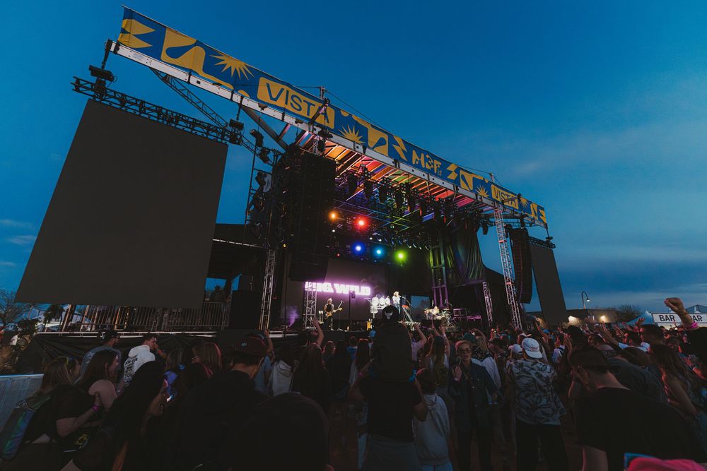 Festival stage with technical equipment and lighting arrays silhouetted against a bright blue sky.