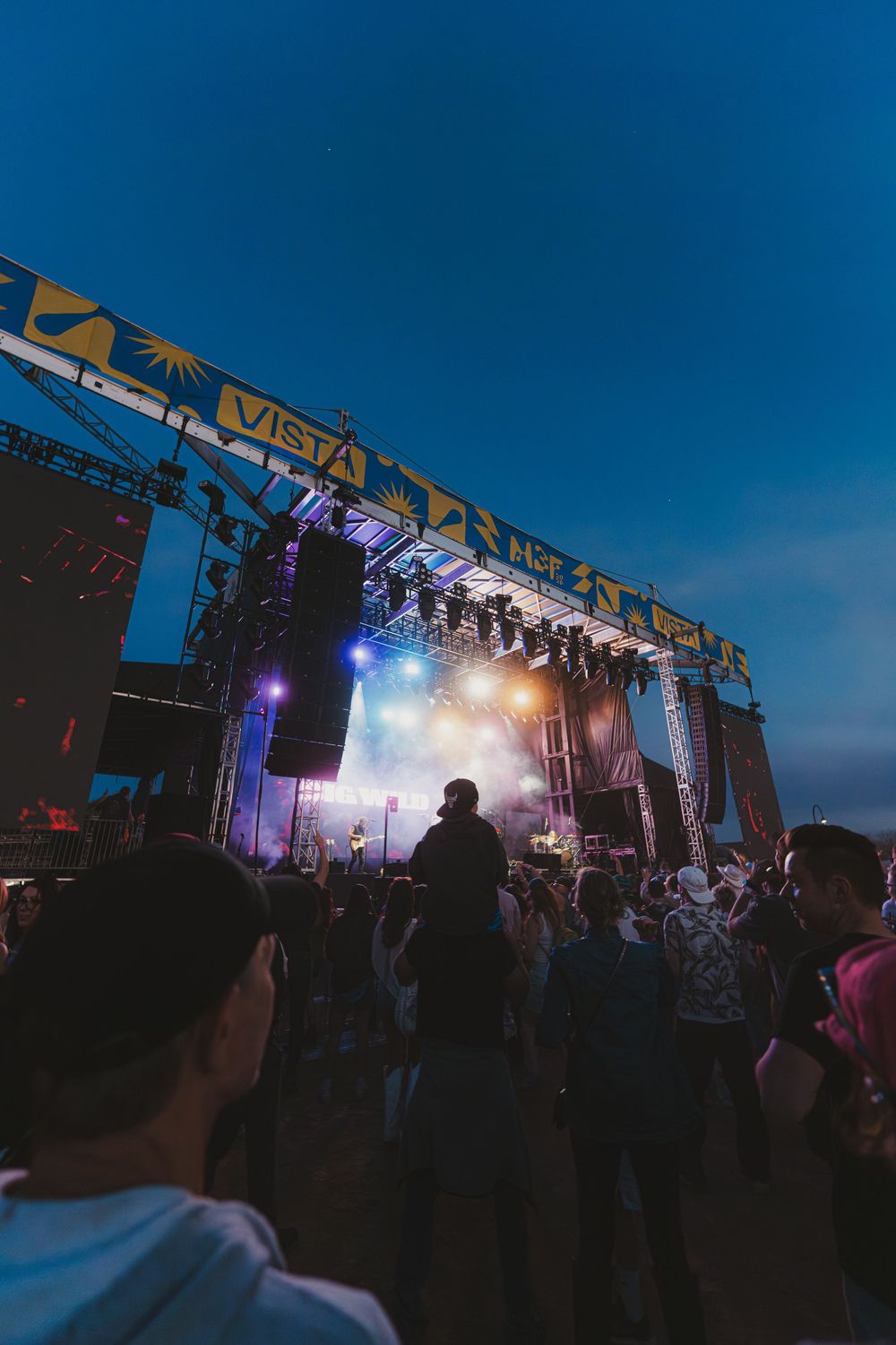 Concert crowd silhouettes gather under colorful stage lights at an outdoor music festival during twilight hours.