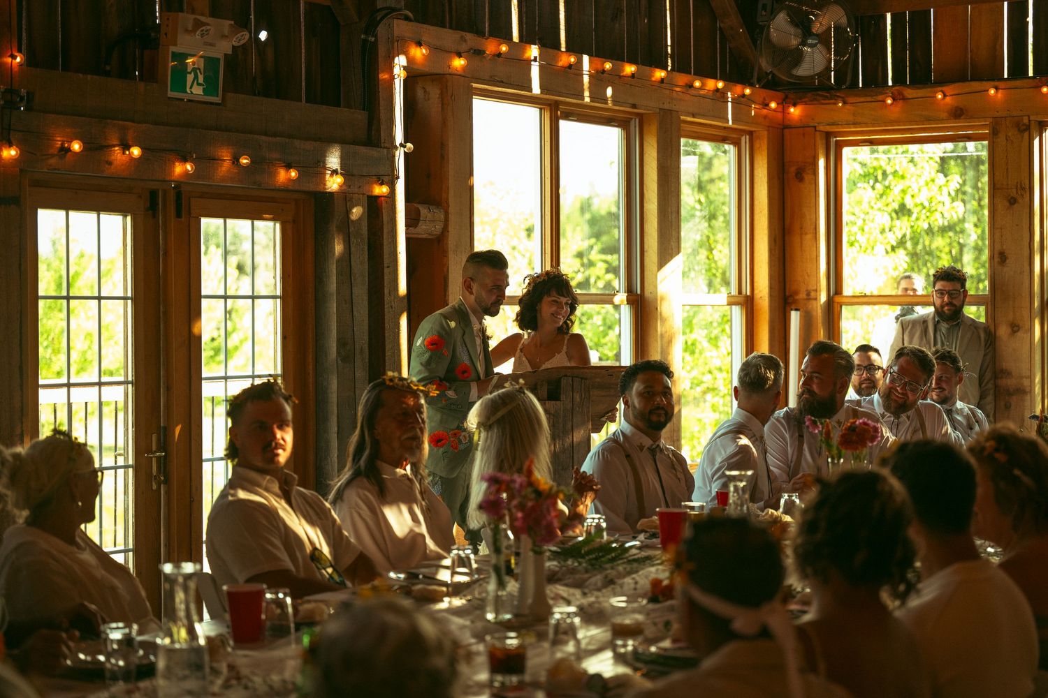 Diners enjoy a meal inside a rustic restaurant with warm lighting and large windows overlooking greenery outside.
