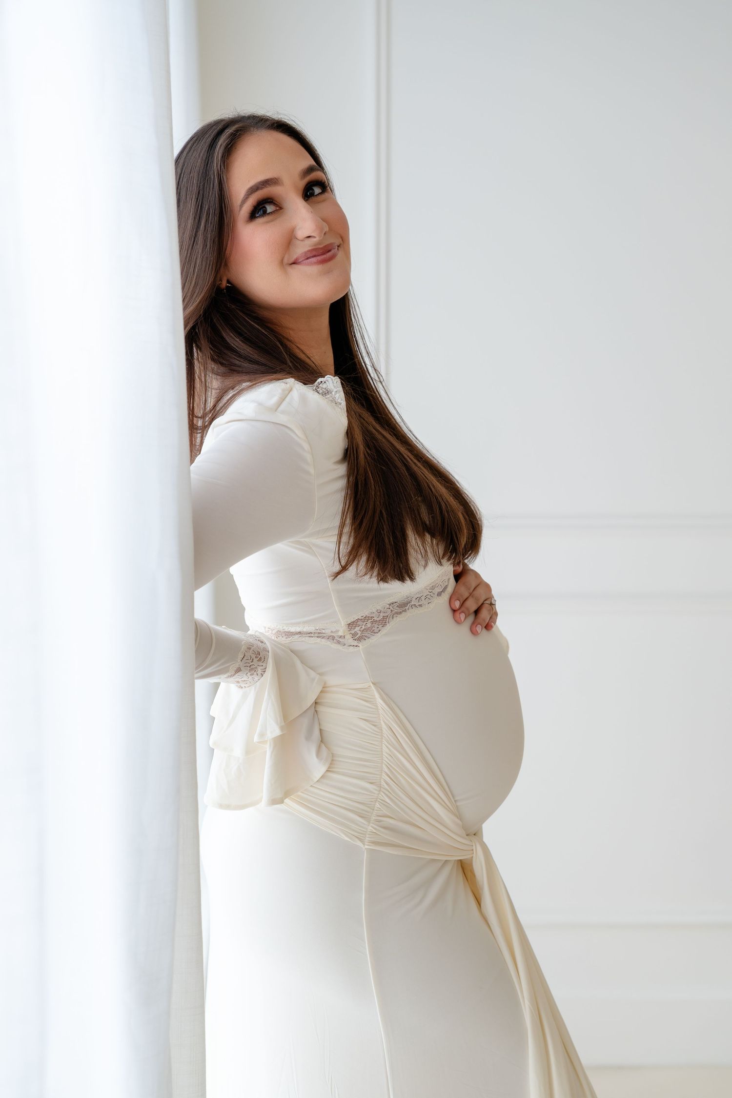 Woman in elegant white dress with bell sleeves poses against light background in artistic maternity photoshoot.