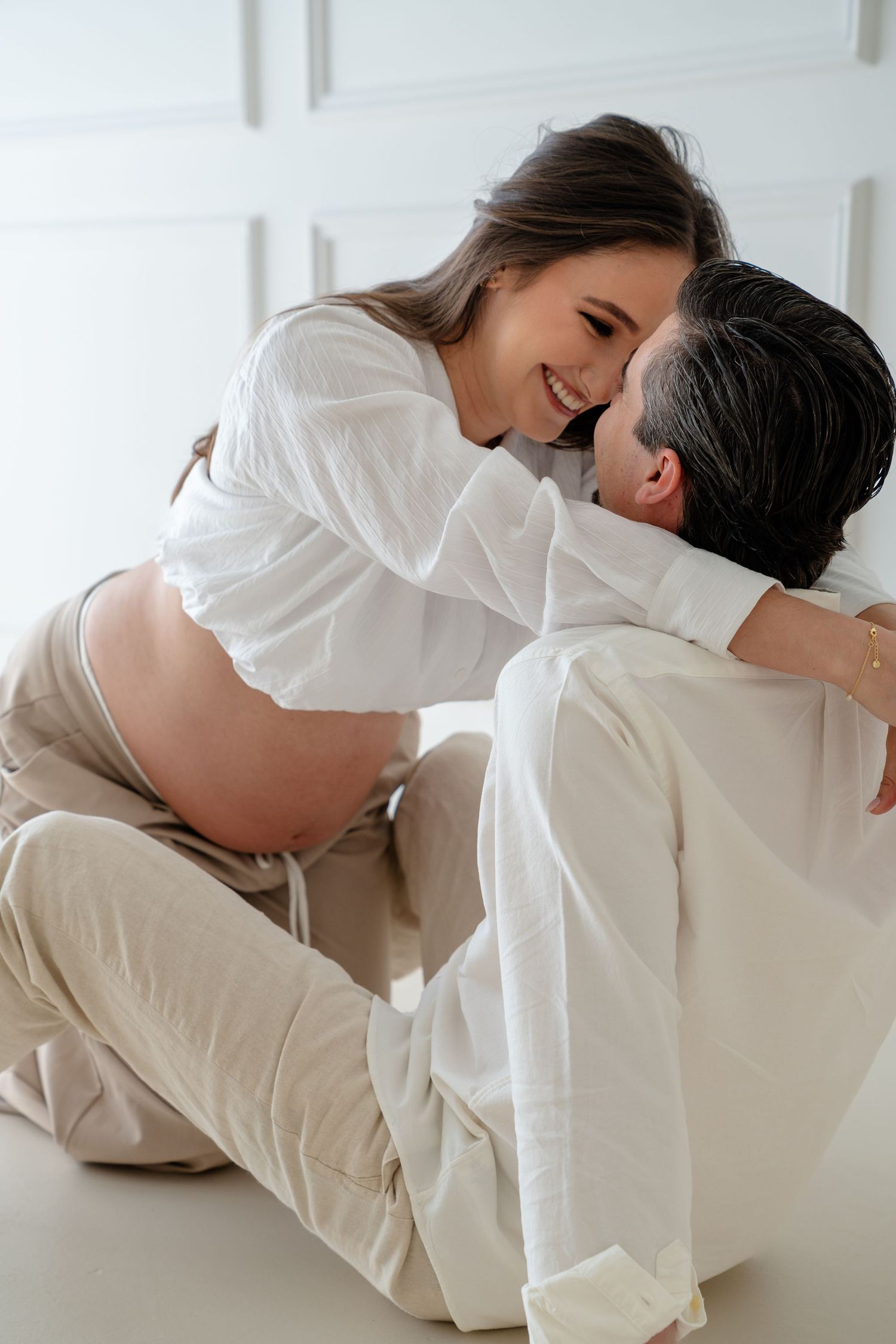 A romantic couple in white clothing embraces tenderly in a bright minimalist setting.