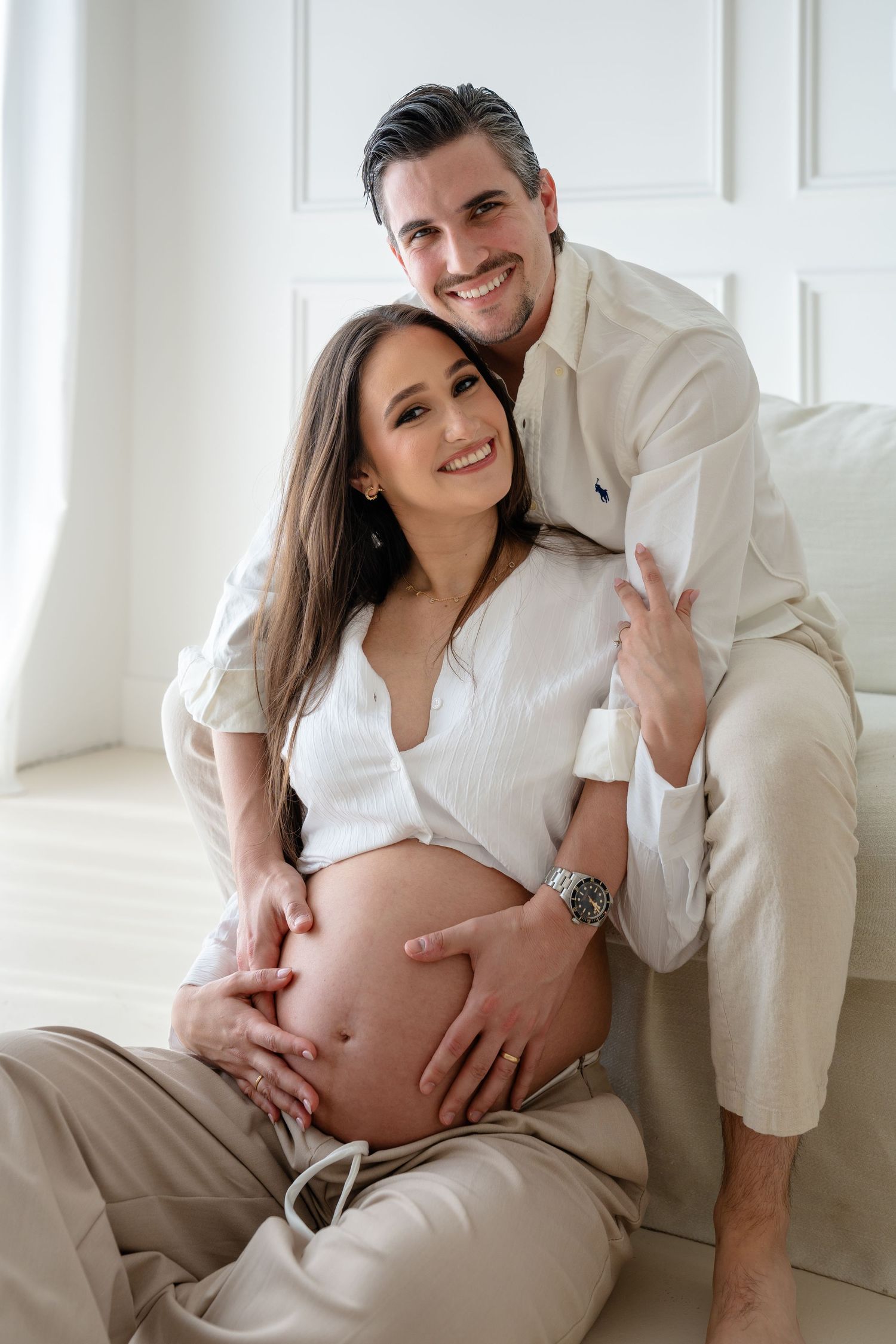 Couple in white clothing poses together for intimate maternity photo session in bright white studio setting.