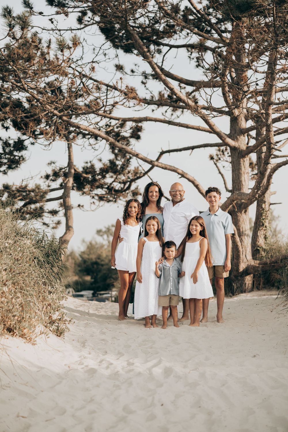 A family group poses together beneath a windswept tree on a sandy beach path during golden hour.