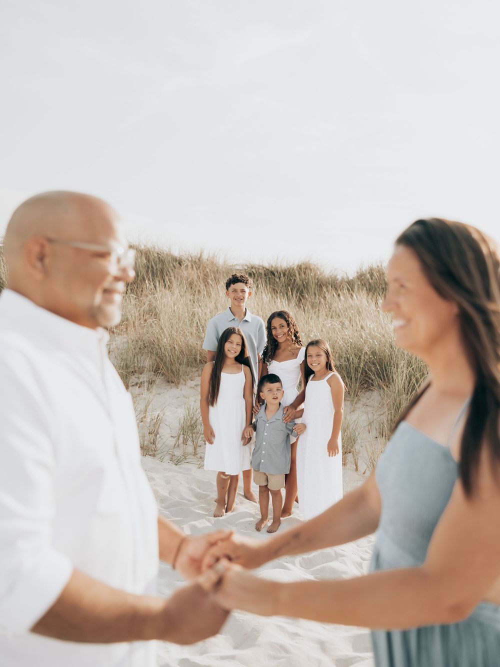 Family wearing coordinated white and light blue outfits poses together on sandy beach at sunset.