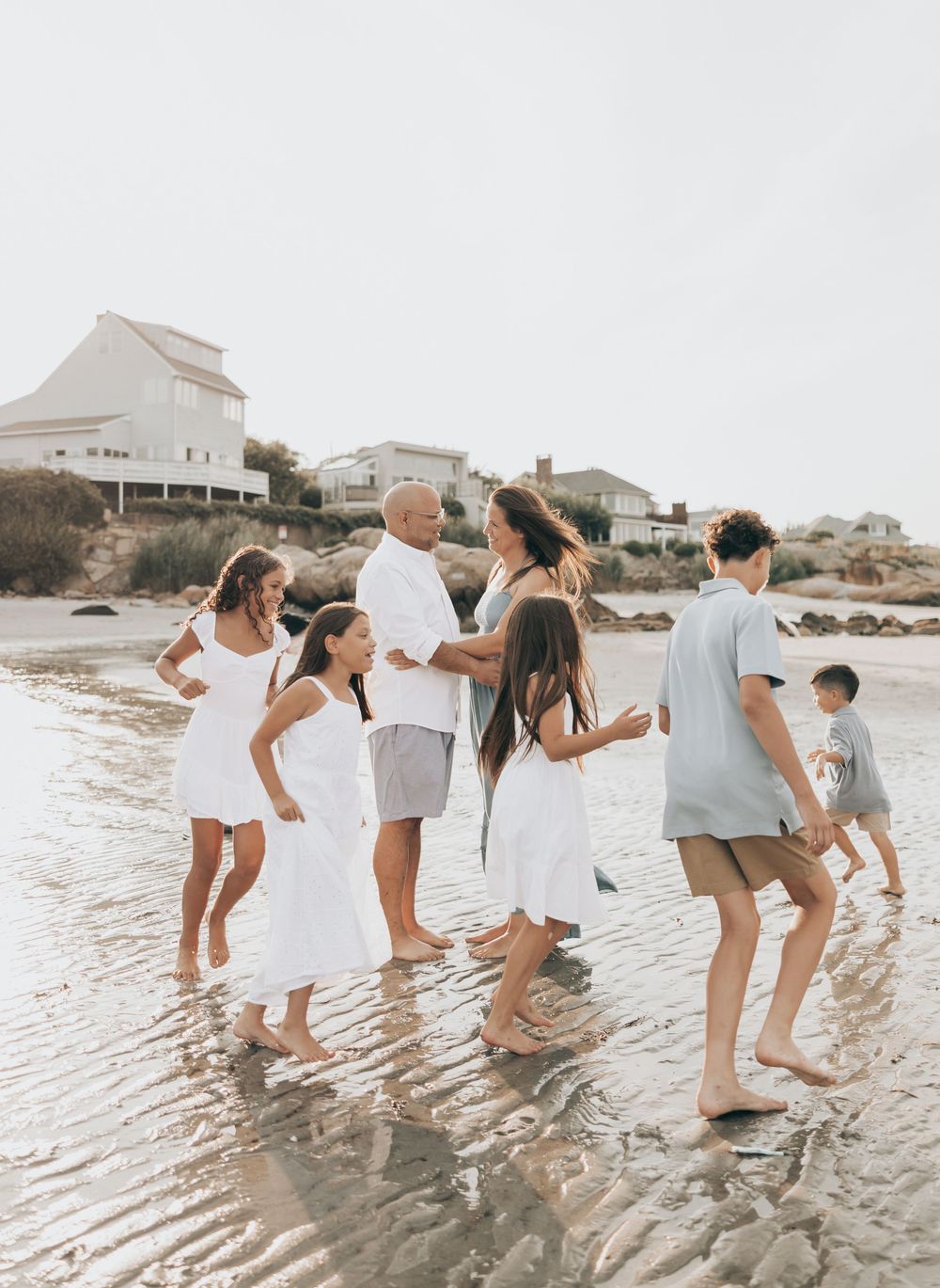 A group of friends celebrating at the beach in white and gray outfits as they dance together in the shallow water.
