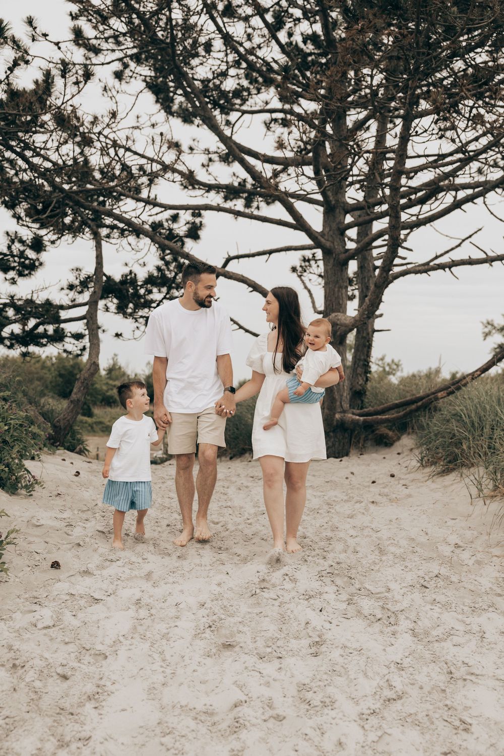 A family of four wearing coordinated white outfits walks together on a sandy beach path beneath towering pine trees.