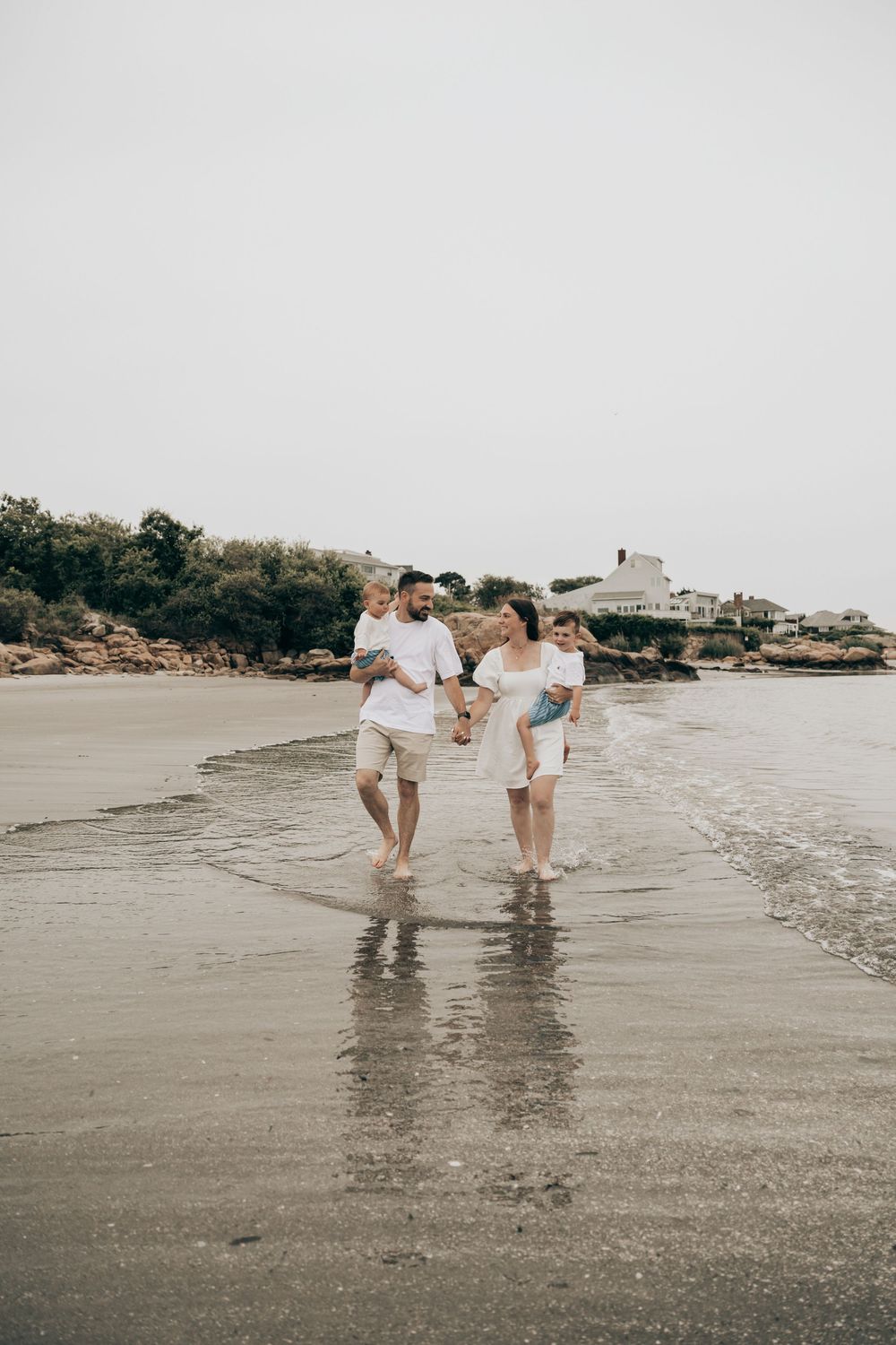 A family strolls together along the wet sand of a beach with coastal homes visible in the background.