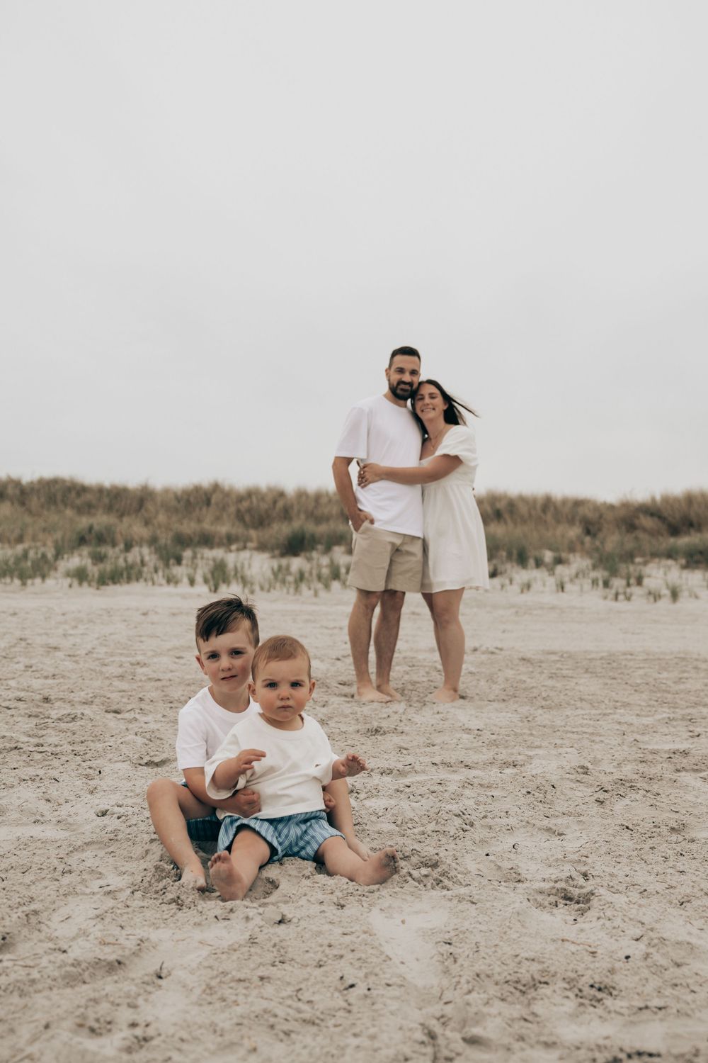A family poses for a beach portrait with parents standing behind while children sit in sand together.