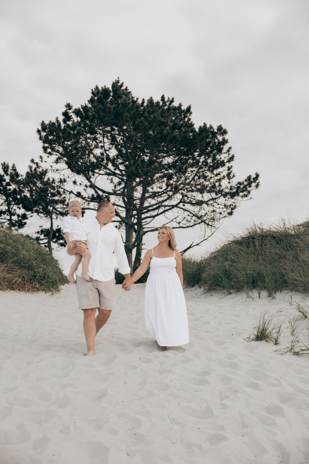 A family in white outfits walks together on a sandy beach with a large pine tree behind them on an overcast day.