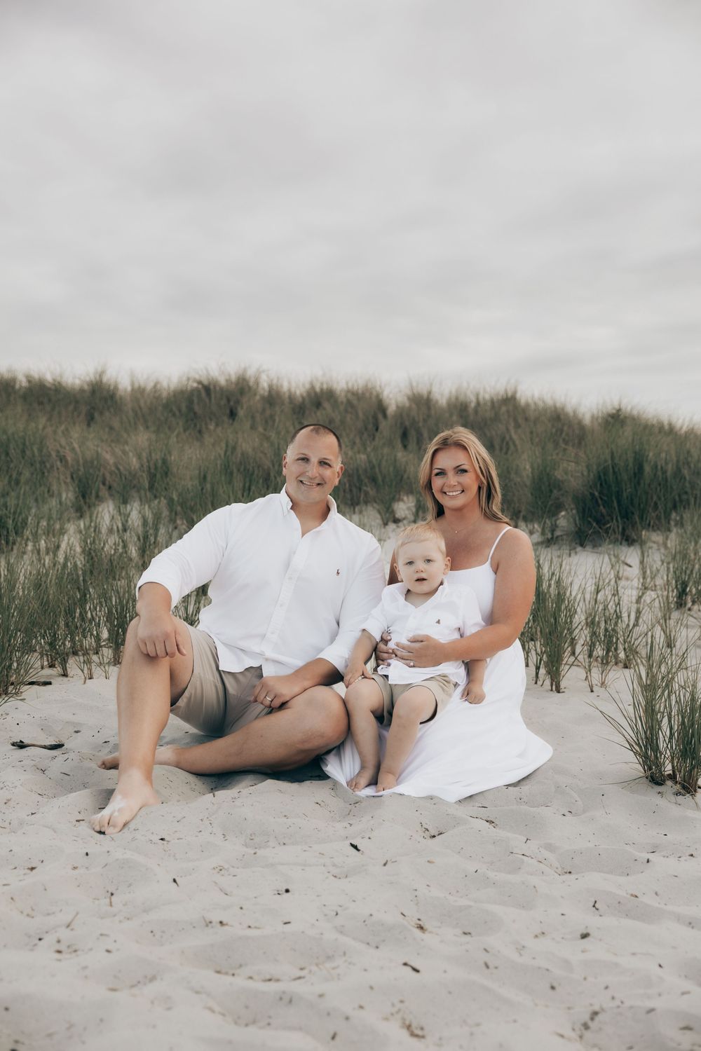 Family in white clothing posed casually on sandy beach dunes with tall grass in the background.