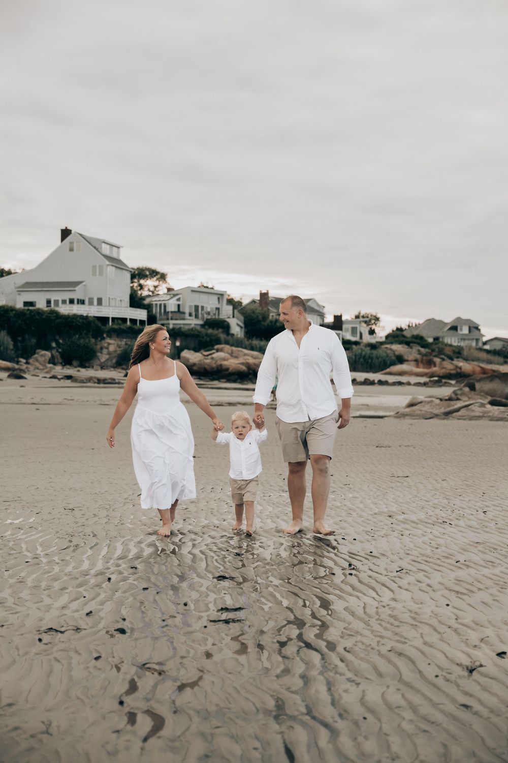 A family in white outfits walks together on a beach with coastal homes in the background on an overcast day.