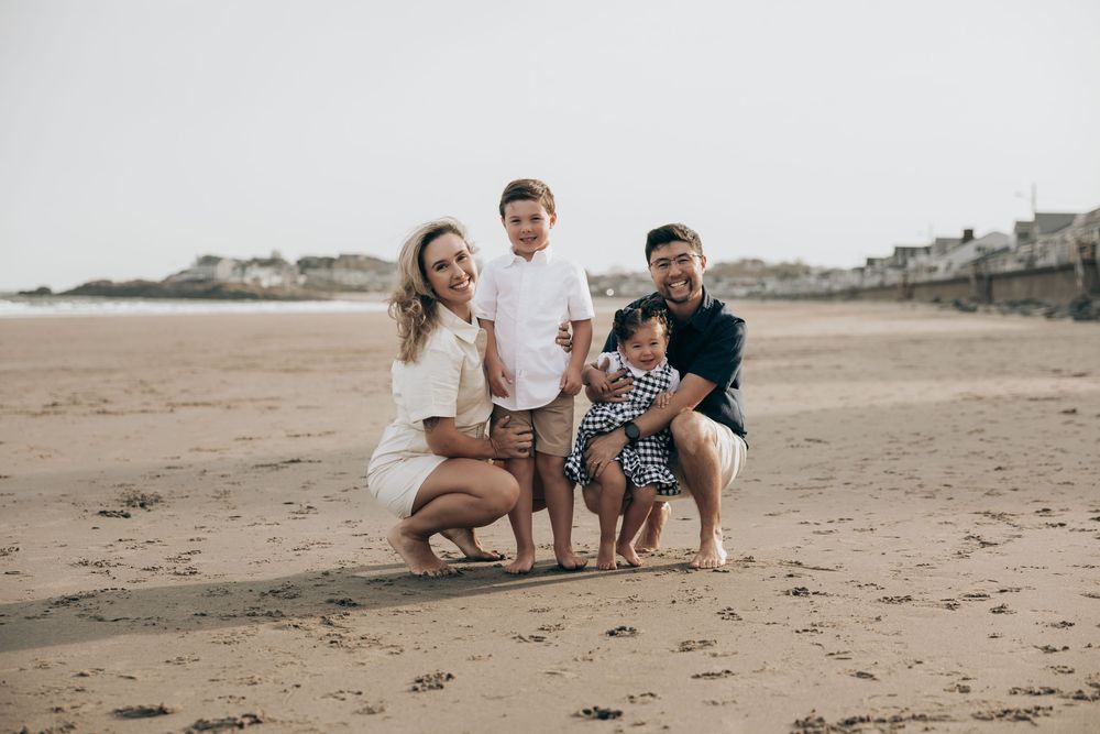 A family of four poses together on a sandy beach at sunset with coastal homes visible in the background.