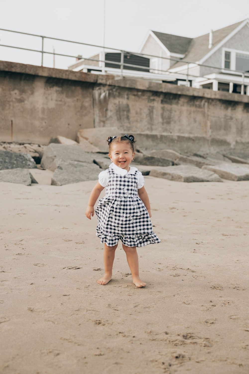 A small child in a black and white gingham dress stands barefoot on a sandy beach near a seawall and coastal house.