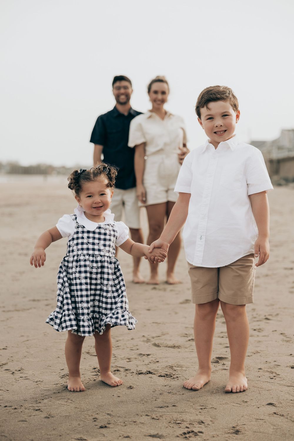 A family stands together at the beach, with two children in the foreground wearing light colored summer clothes.