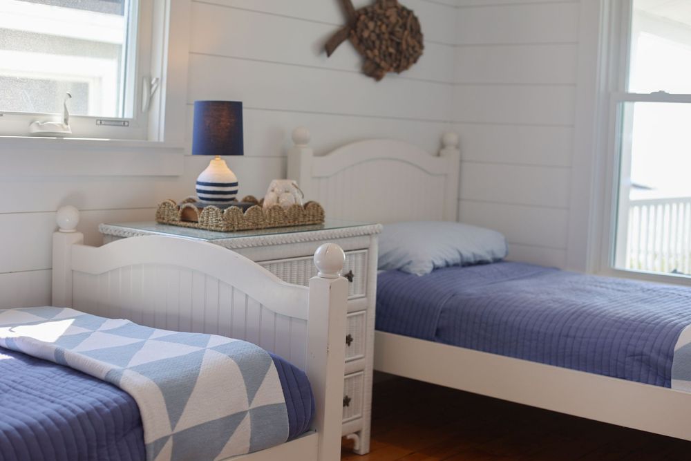 A coastal bedroom with twin beds featuring blue bedding and white furniture in a beach cottage style.