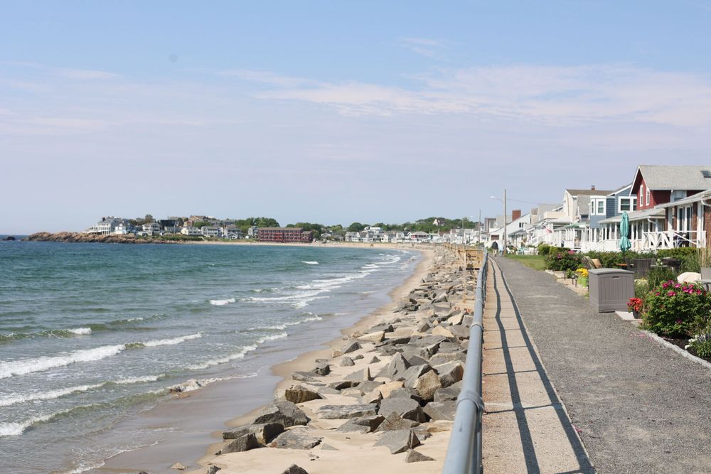 Oceanfront homes line a rocky beach with a paved walkway along the New England coast.