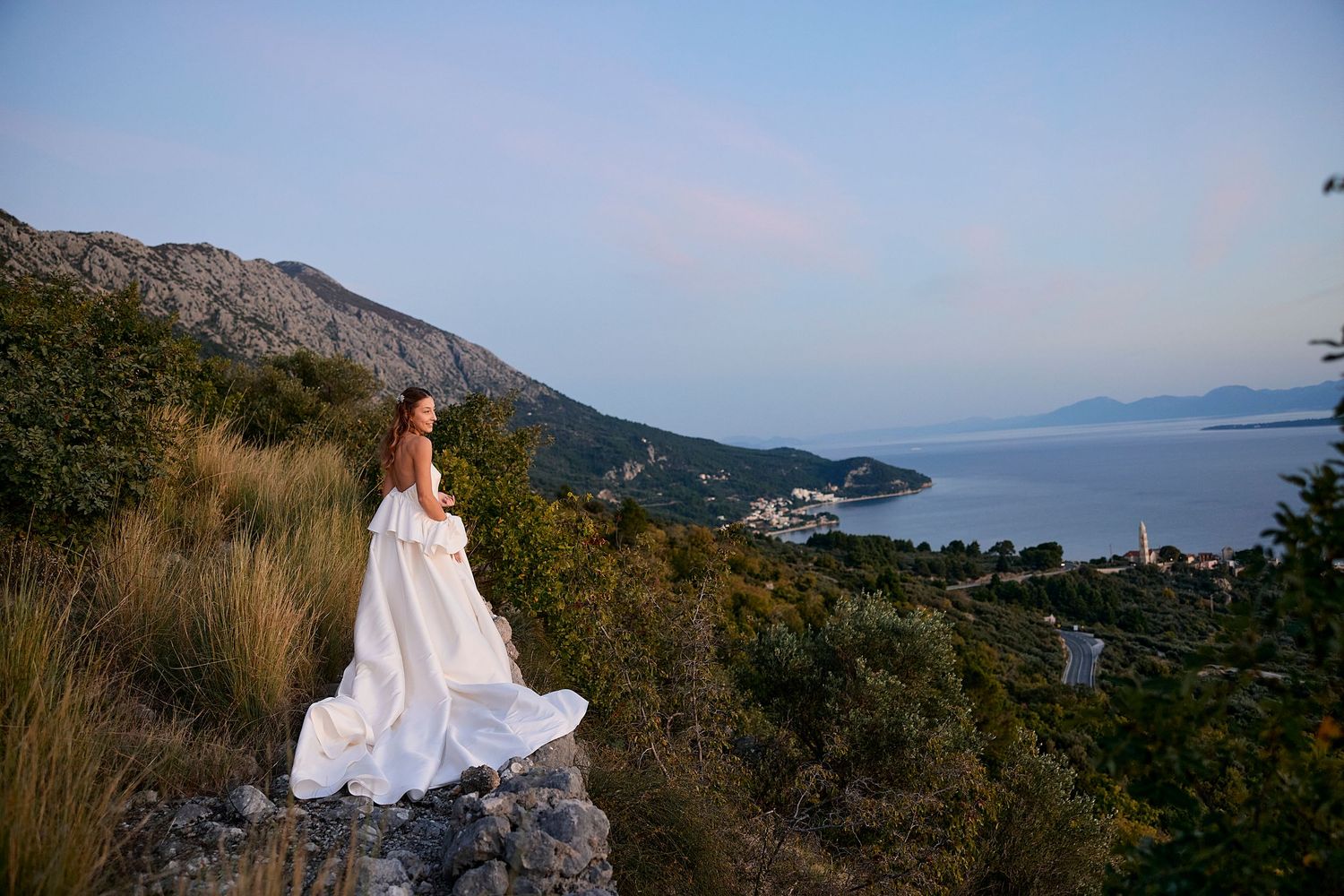 White dress flowing in the wind against a scenic coastal mountain landscape at sunset.