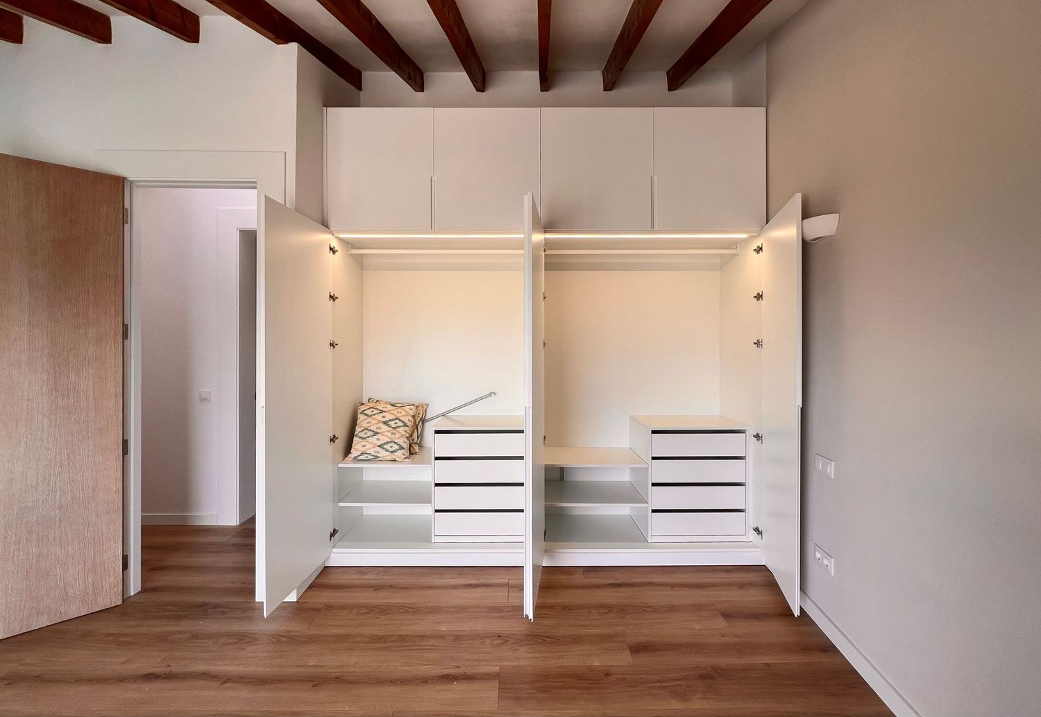 Interior view of a modern built-in wardrobe with white drawers and hanging rails installed in a room with wooden floors.