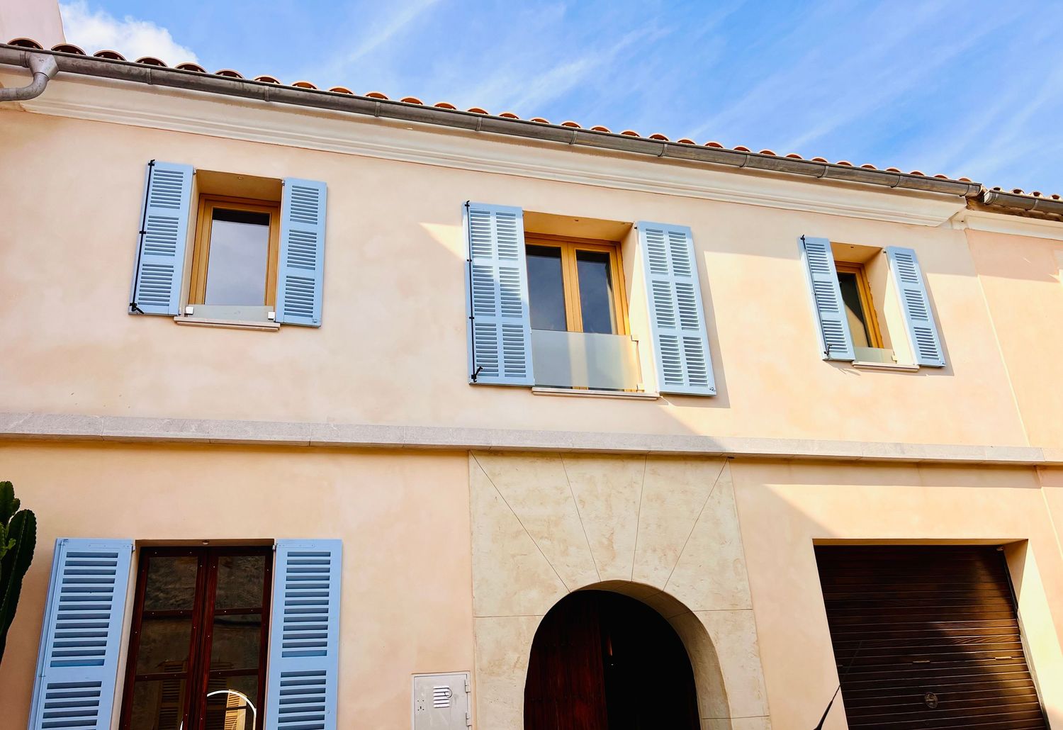 Mediterranean style house exterior with pale yellow walls and blue shuttered windows under bright sunlight.
