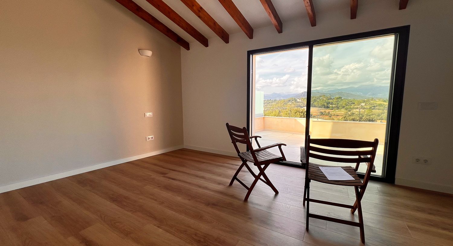 Minimalist room with wooden floors, exposed beams, and two chairs facing a large window with mountain views.