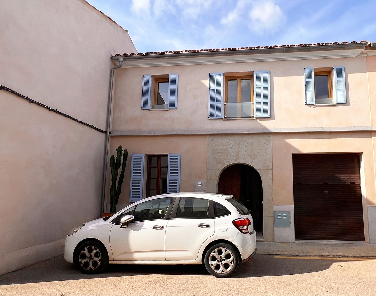 White car parked in front of beige Mediterranean style house with blue shutters and arched entrance.