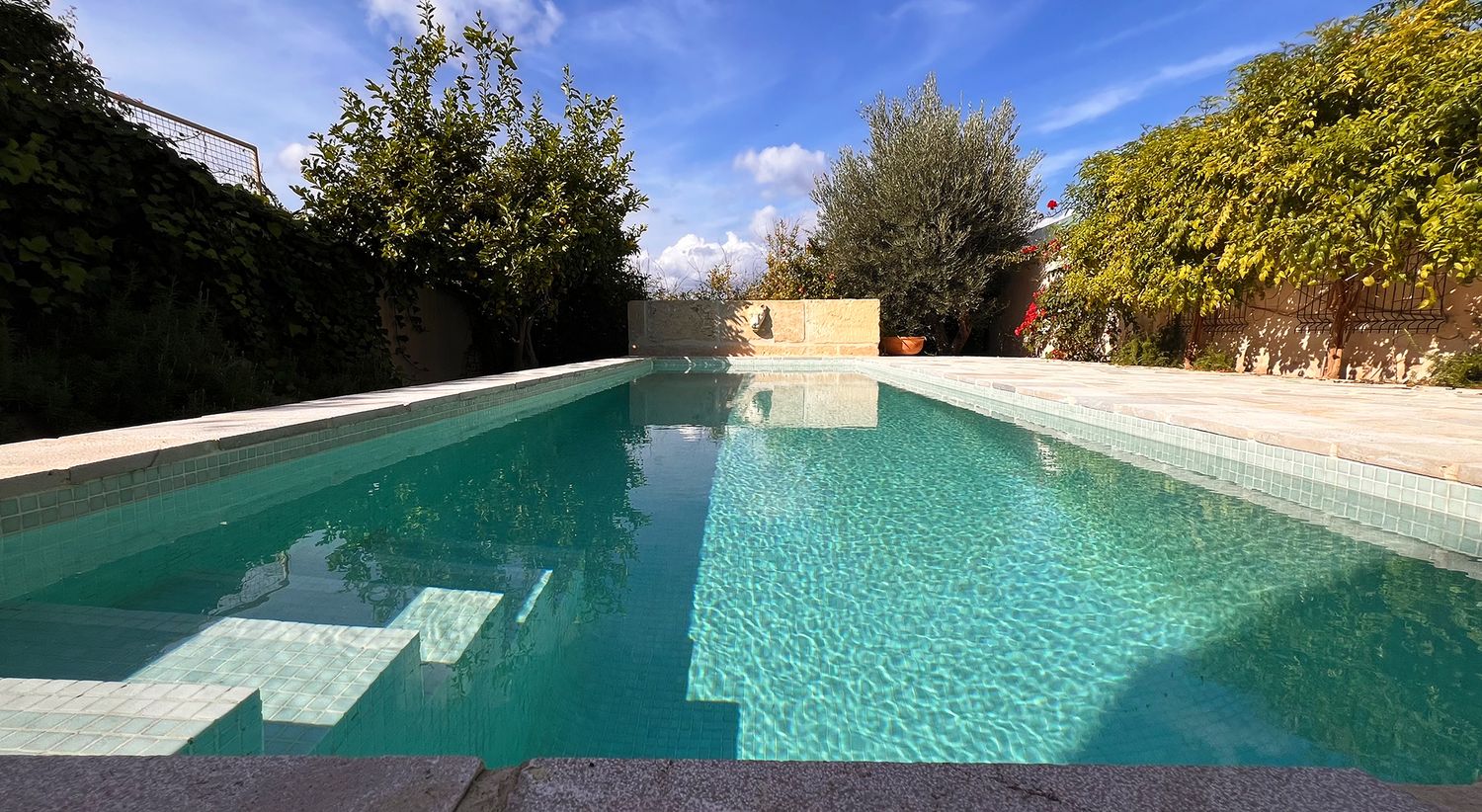 Rectangular swimming pool with turquoise water surrounded by stone walls and lush garden landscaping.