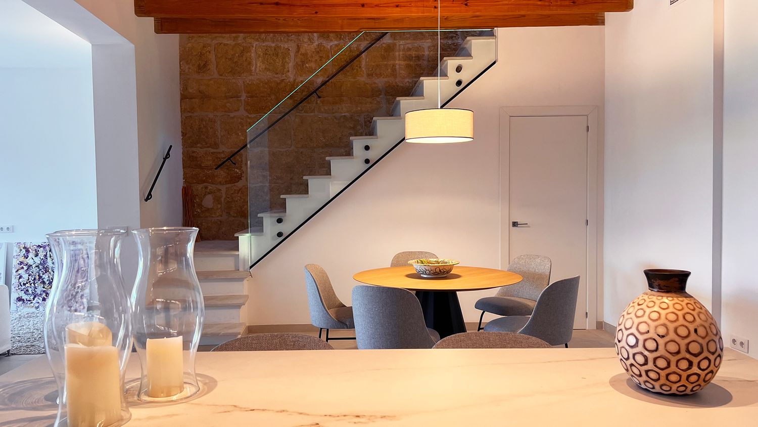 Contemporary dining area under a wooden beam ceiling with glass staircase and decorative vase.