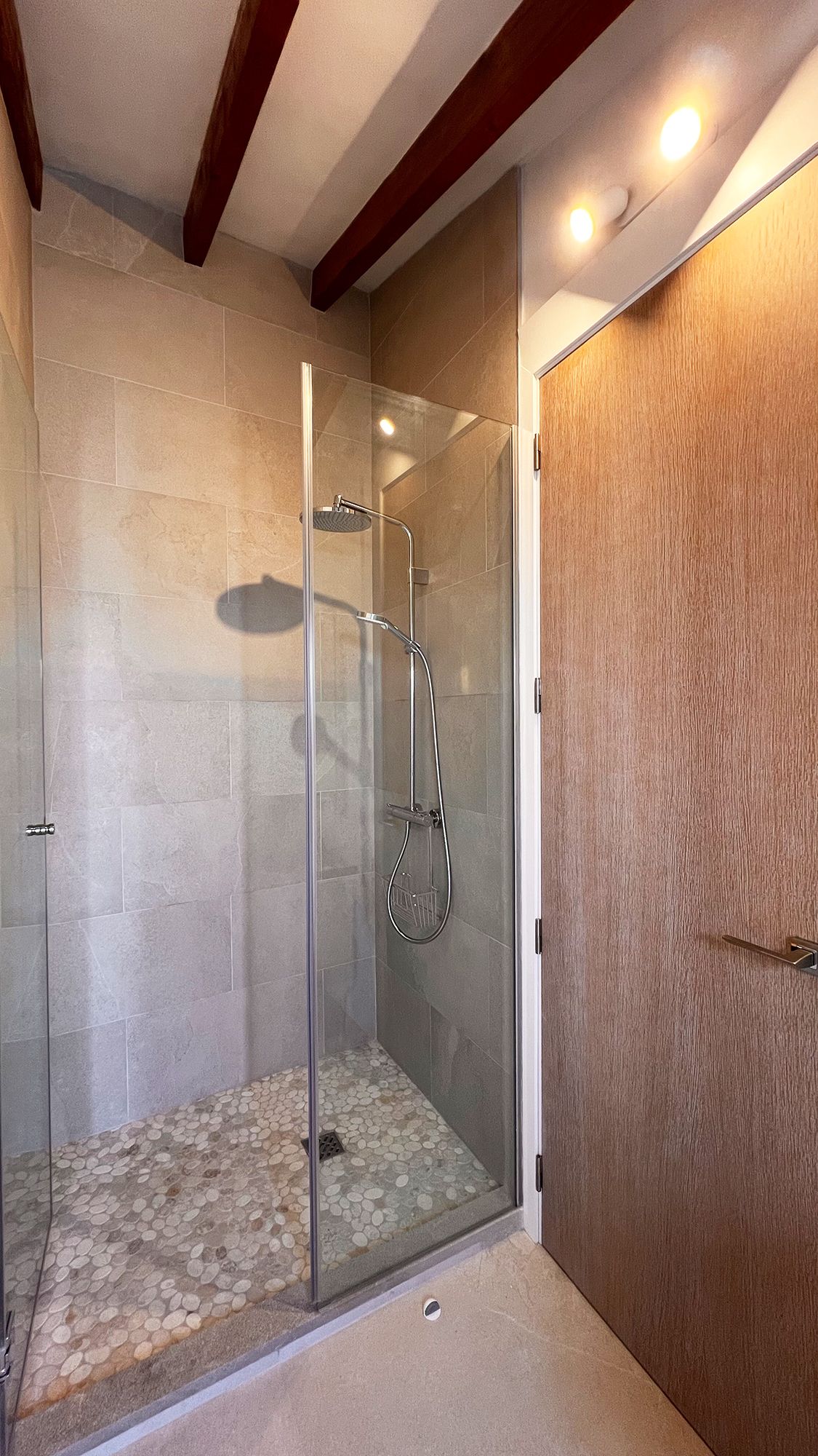 Modern shower enclosure with glass doors and pebble floor under wooden beams.