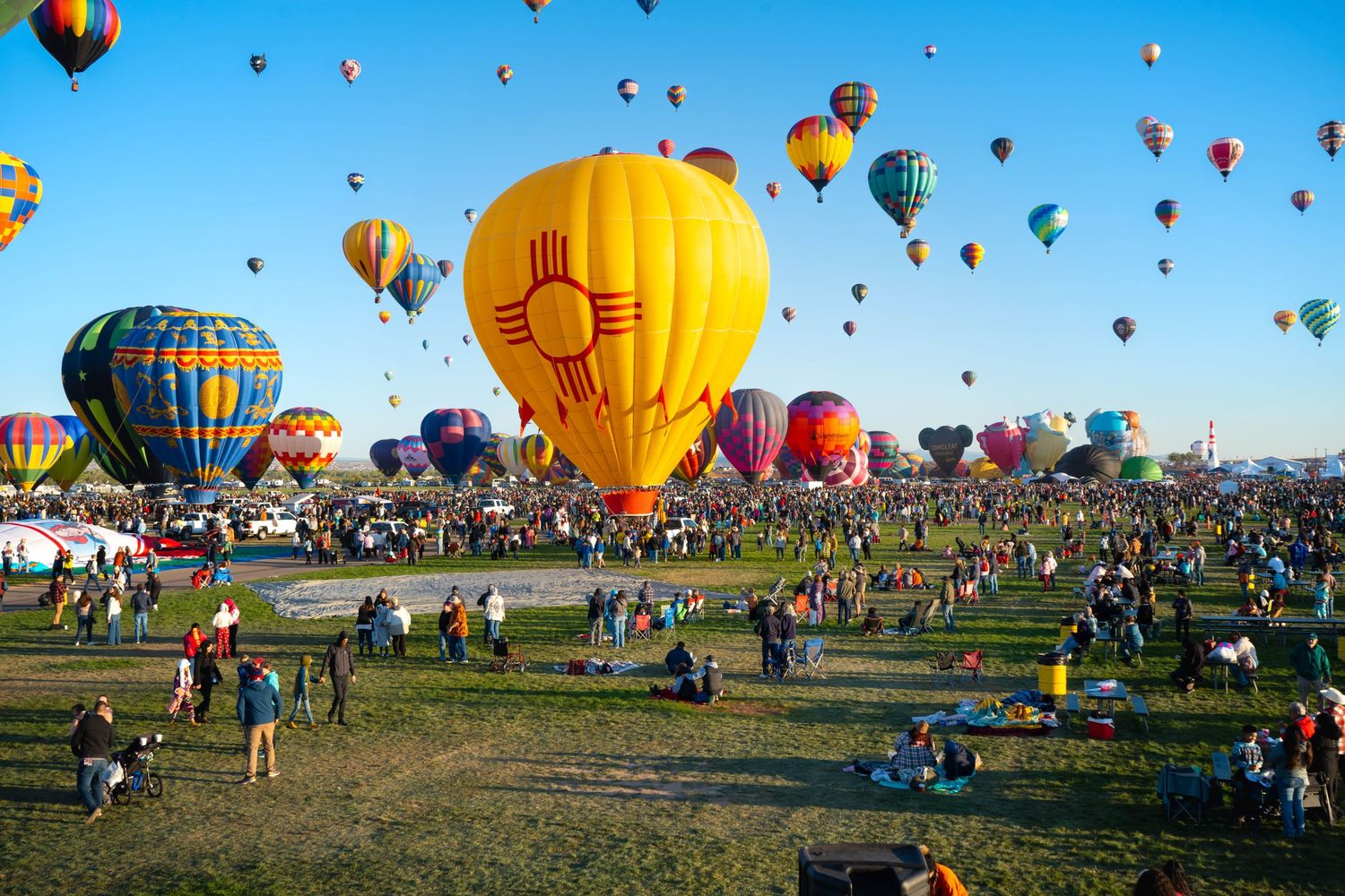 Hot air balloons ascending at Albuquerque Balloon Fiesta with yellow Zia balloon in foreground.