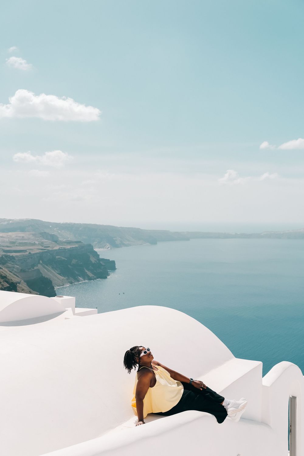 Woman in yellow top posing on white Santorini rooftop with stunning Aegean Sea caldera views on sunny day.