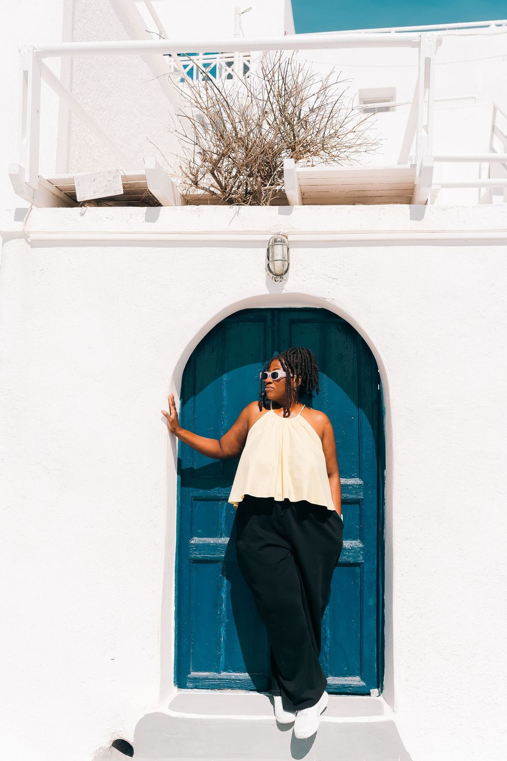 Woman in yellow top and black pants posing by a blue arched door on a white Grecian building.