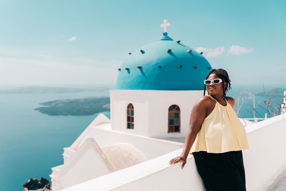 Woman in yellow top posing by iconic blue-domed church in Santorini, Greece, with caldera views.