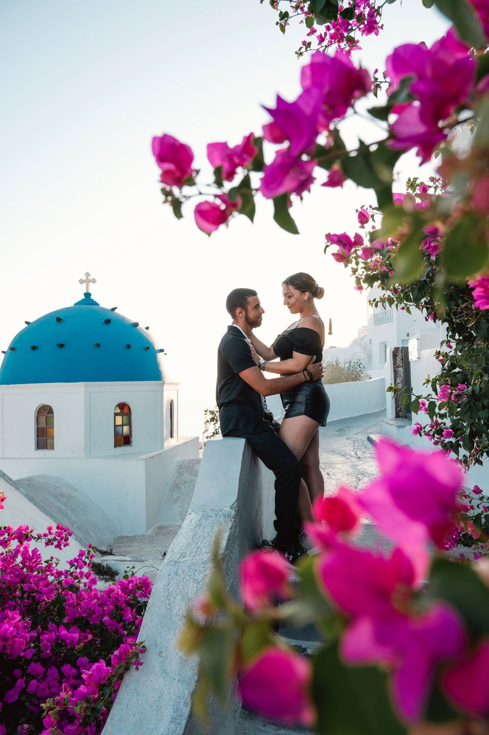 Intimate couple portrait framed by vibrant bougainvillea flowers and blue-domed church in Santorini.