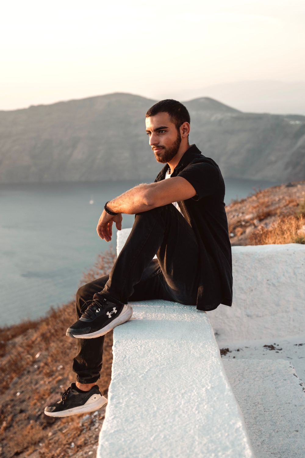 Person in black clothing sitting on white wall overlooking Mediterranean cliffs at sunset.