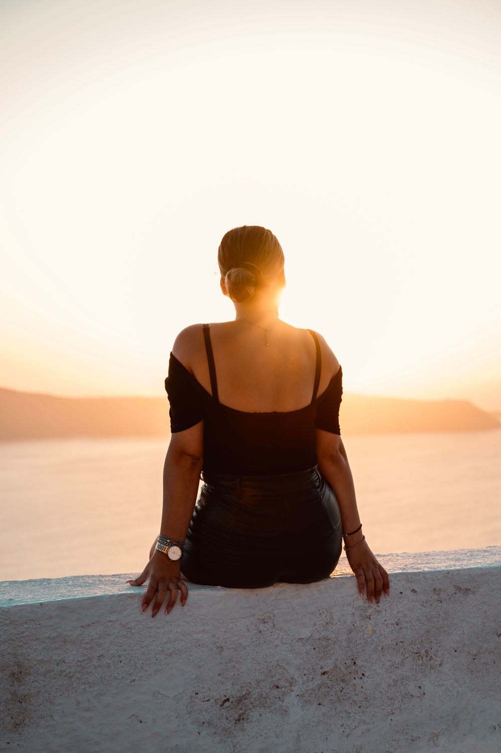 Person sitting on white ledge watching sunset over calm waters and mountains in Greece.