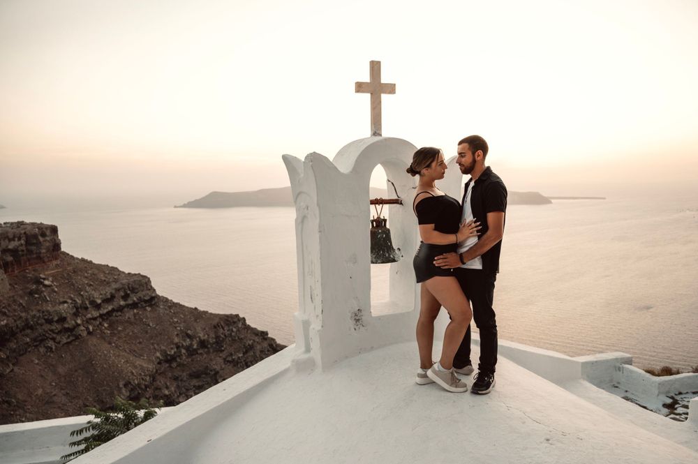 Couple standing by traditional Greek church with white dome and cross overlooking the Aegean Sea.
