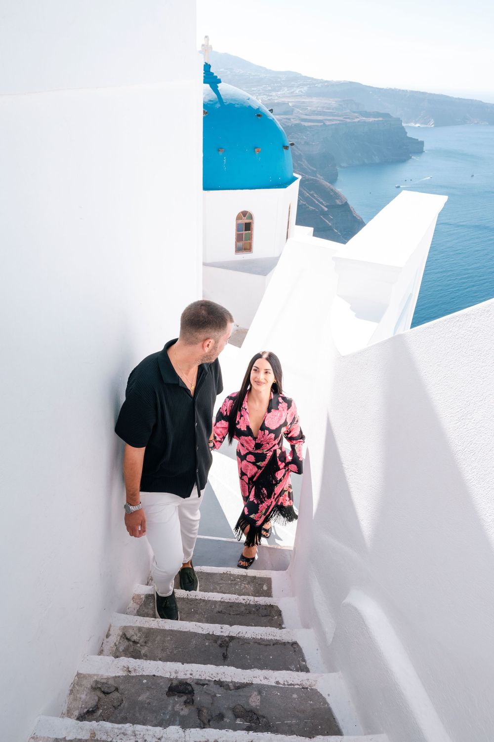 Couple on white Santorini steps with iconic blue domed church in background.
