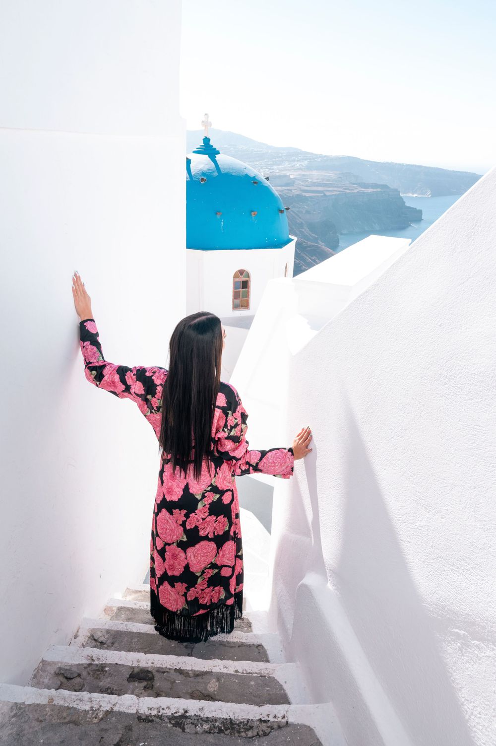 Person in floral dress walking down white steps with blue domed church in Santorini.